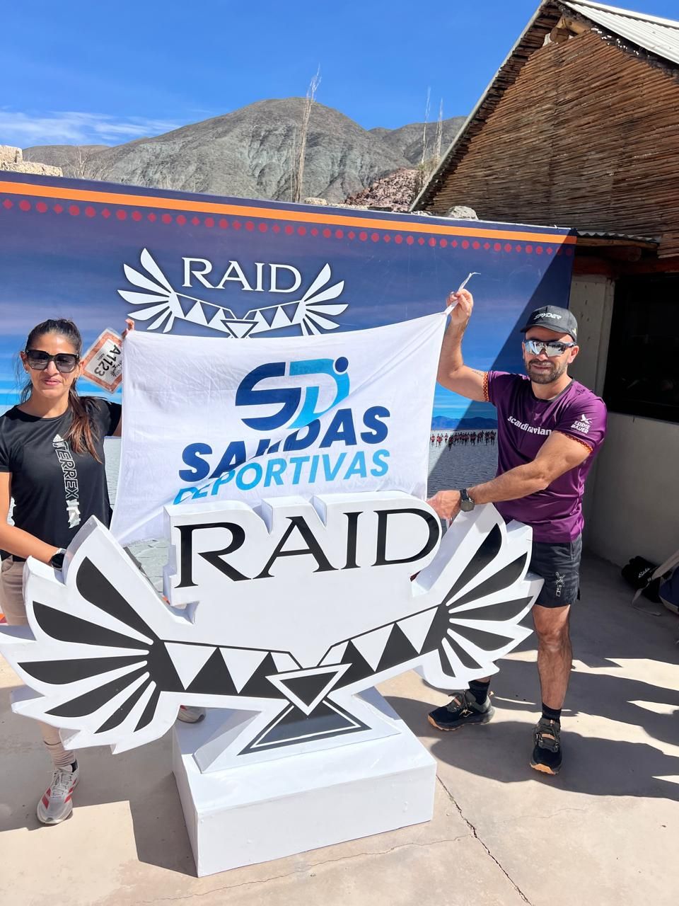 Dos corredores con la bandera de Salidas Deportivas junto al cartel del Raid